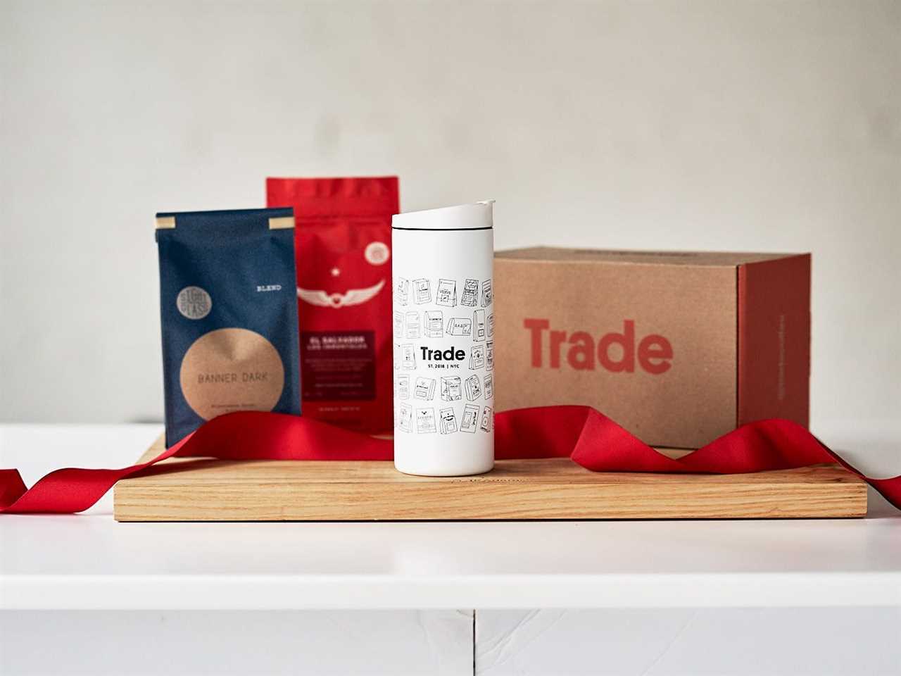 a red ribbon runs between bags of coffee, a thermos, and a box with Trade coffee’s logo on it as part of the best coffee subscription in 2021