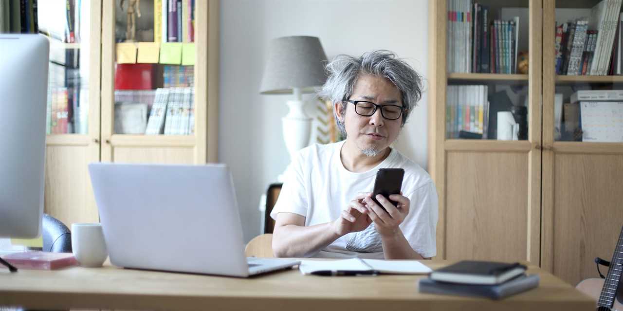 Man texting on phone in home office next to laptop