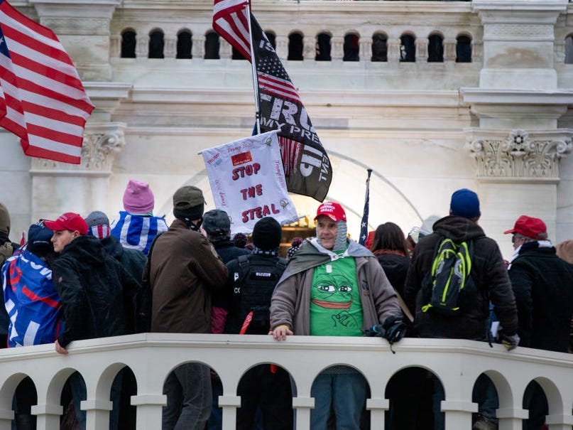 A man wears a red Make America Great Again hat and a "Groyper" meme shirt showing the white nationalist group's Pepe the Frog-inspired logo. He is surrounded by pro-Trump protesters waving America and Trump flags outside the US Capitol building.