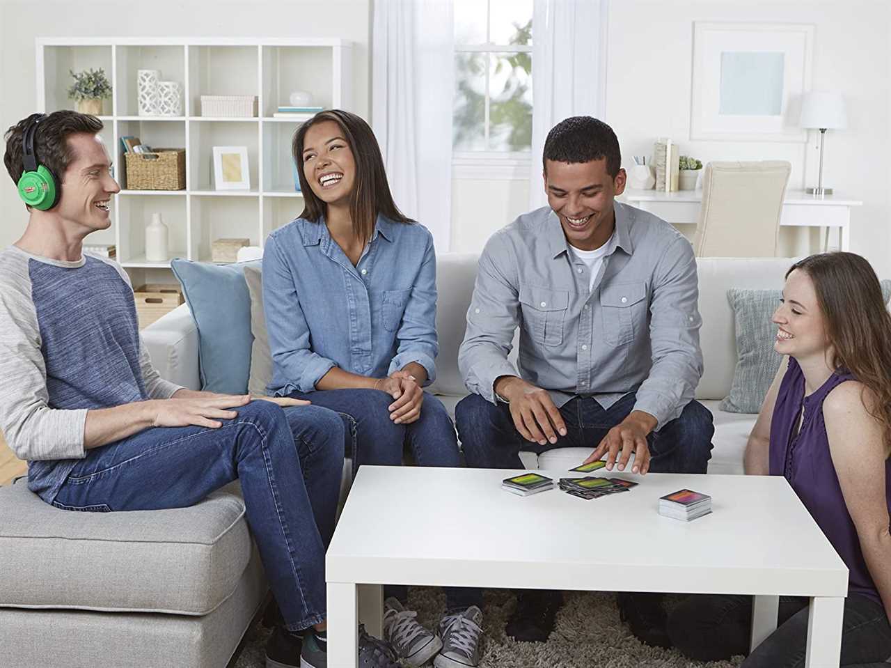 A photo of a group of friends sitting on a gray couch, one is wearing headphones, and a deck of cards is on the table