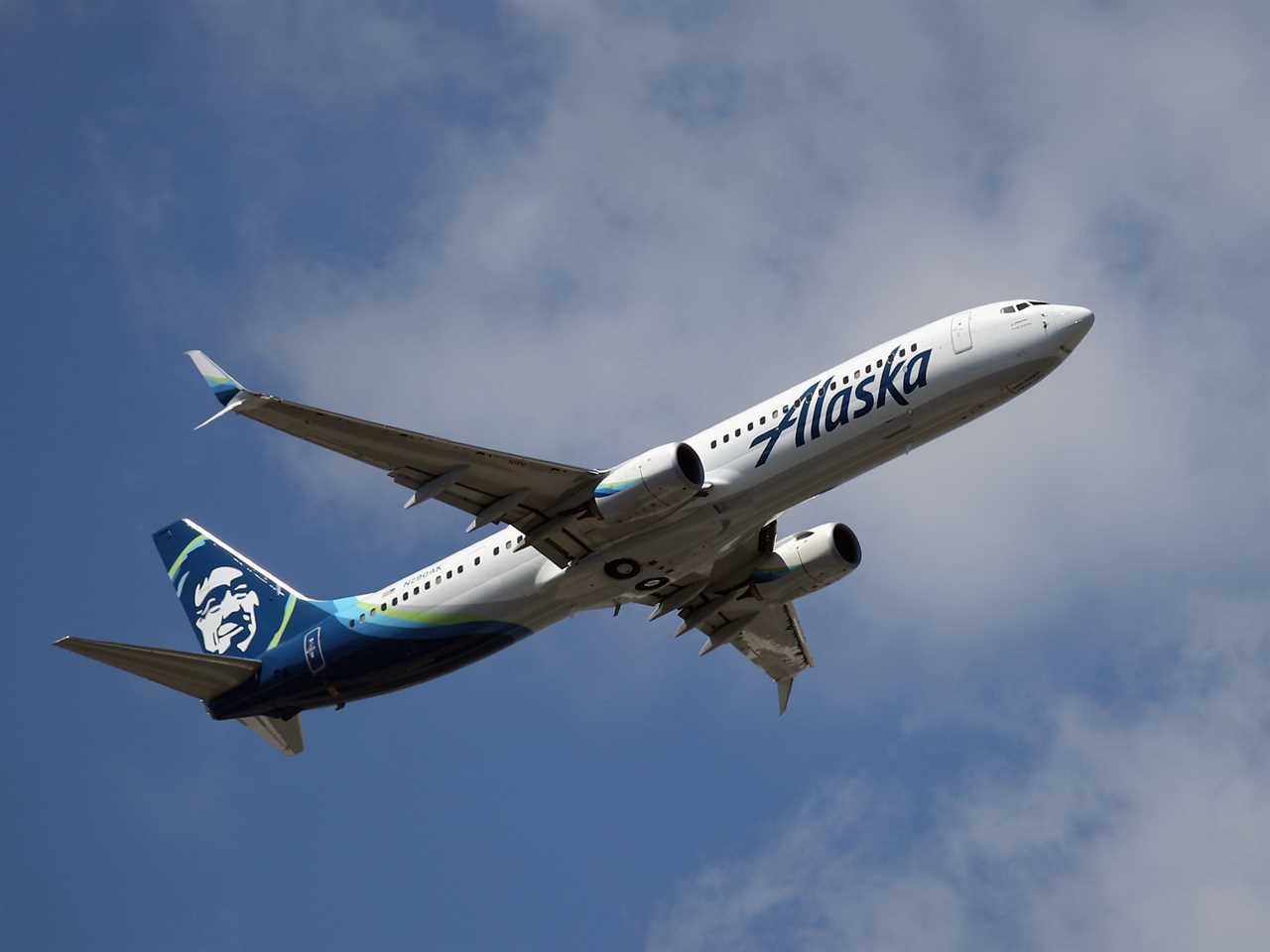 An Alaska Airlines airplane taking off against a blue sky with white clouds.