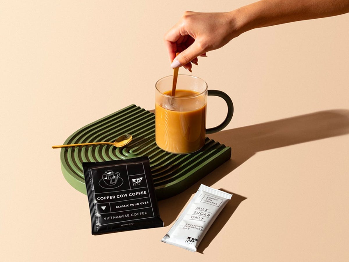 Hand stirring a glass cup of coffee behind Copper Cow Coffee filter and condensed milk - Best asian american food and drink brands in 2021