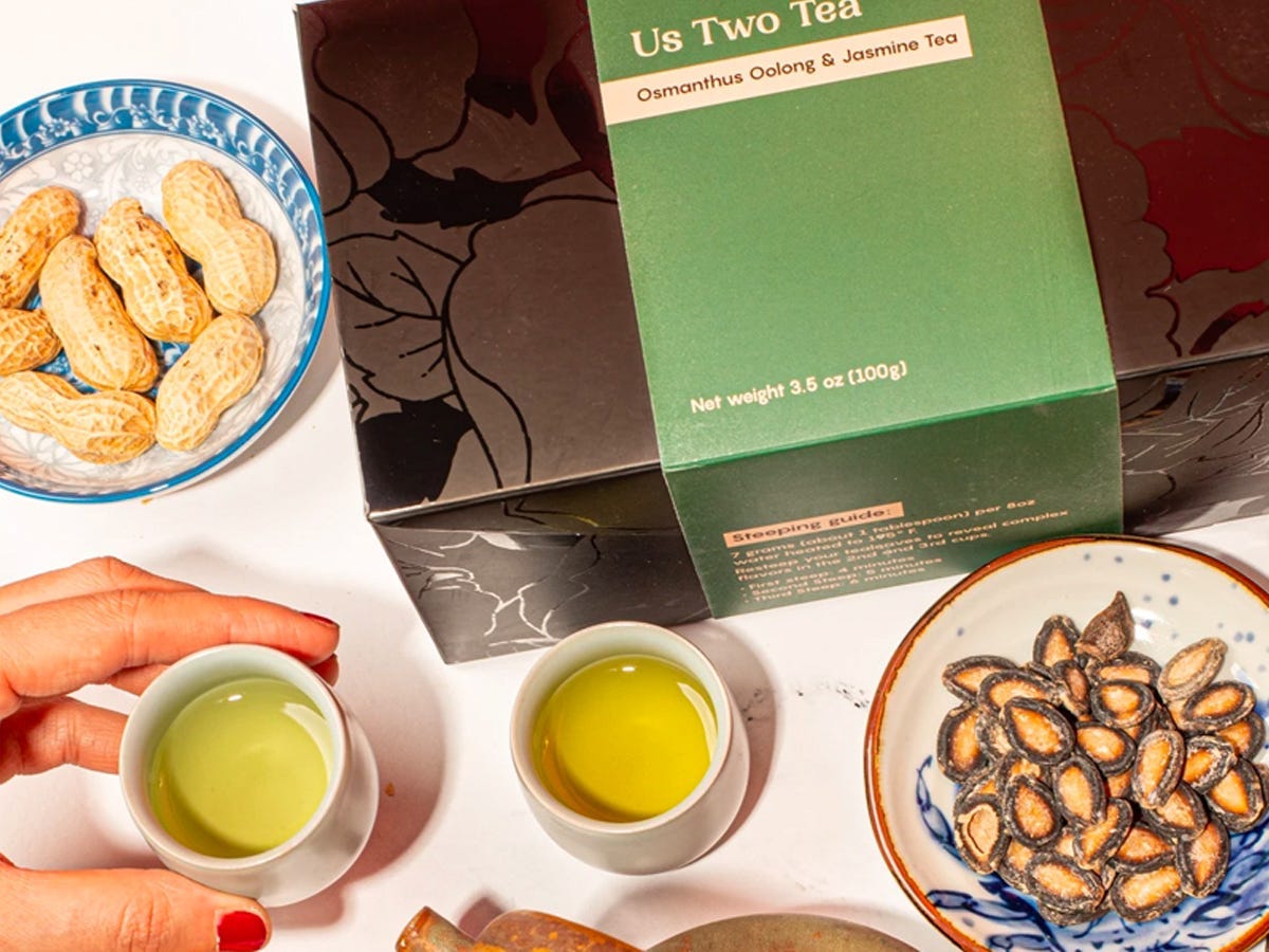 Hand reaching for small cup of jasmine tea from Us Two Tea on a table with peanuts and watermelon seeds part - Best Asian American food brands in 2021