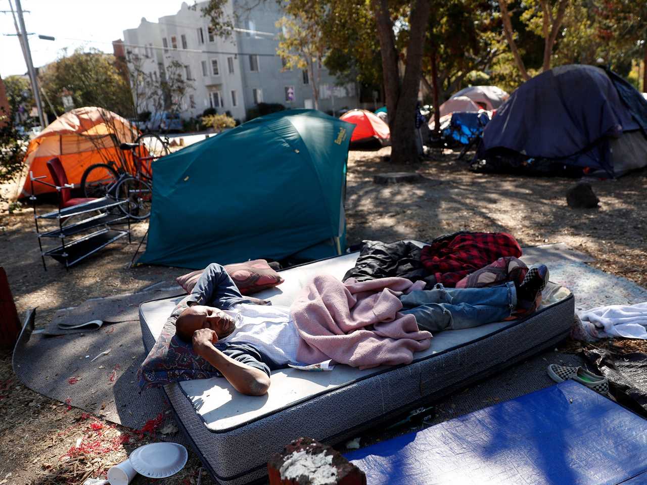 A man lays on a mattress in a park surrounded by tents.