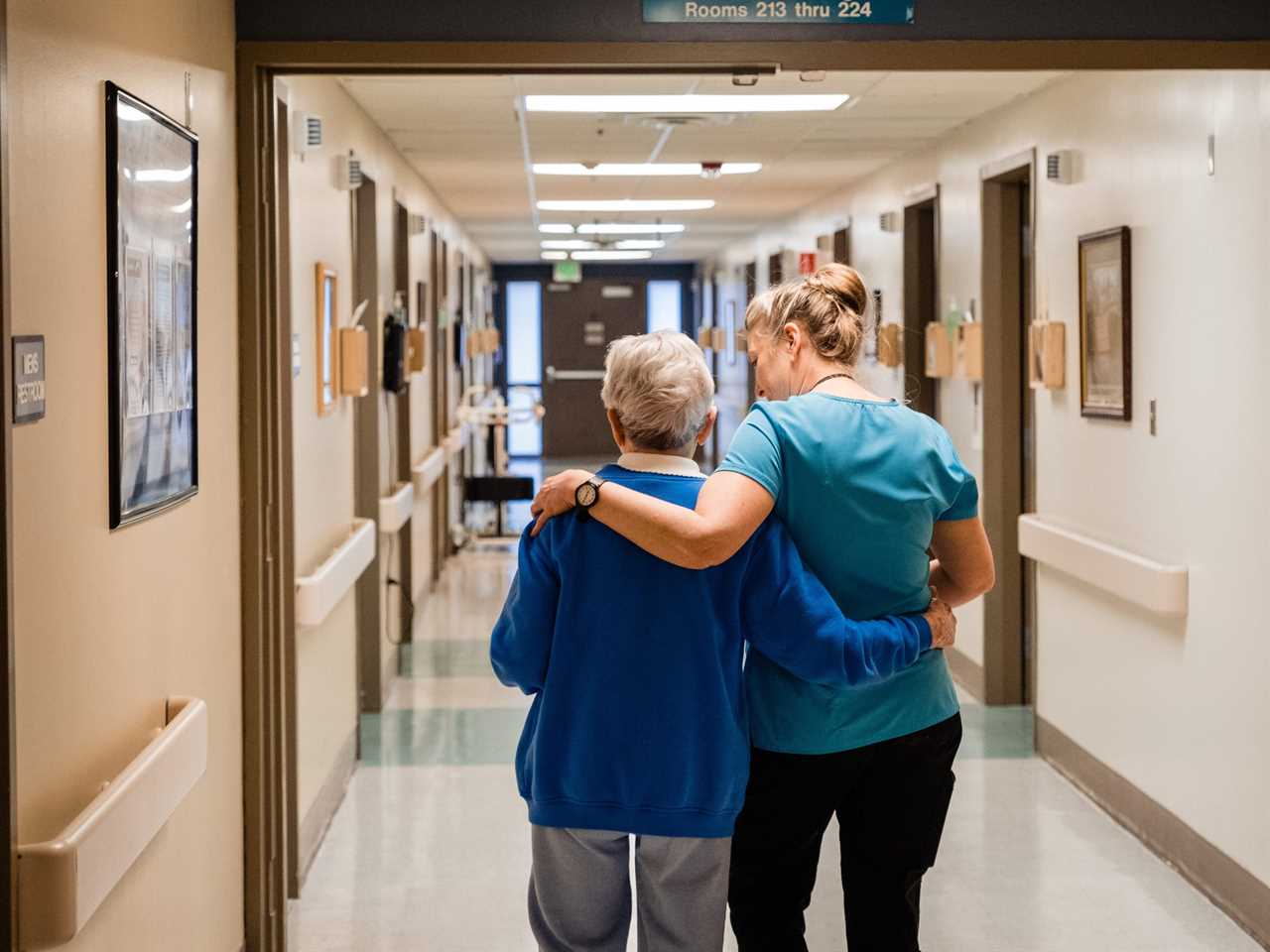 A female nurse has her arm around an elderly woman in a blue sweater. They are walking down a hallway in a hospital.