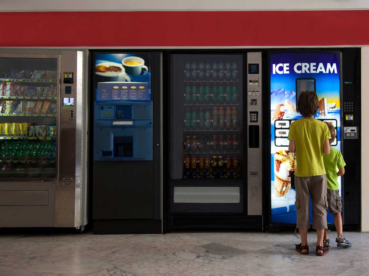 Two children standing in front of an ice cream vending machine. There are three other kinds of vending machines in the photo.