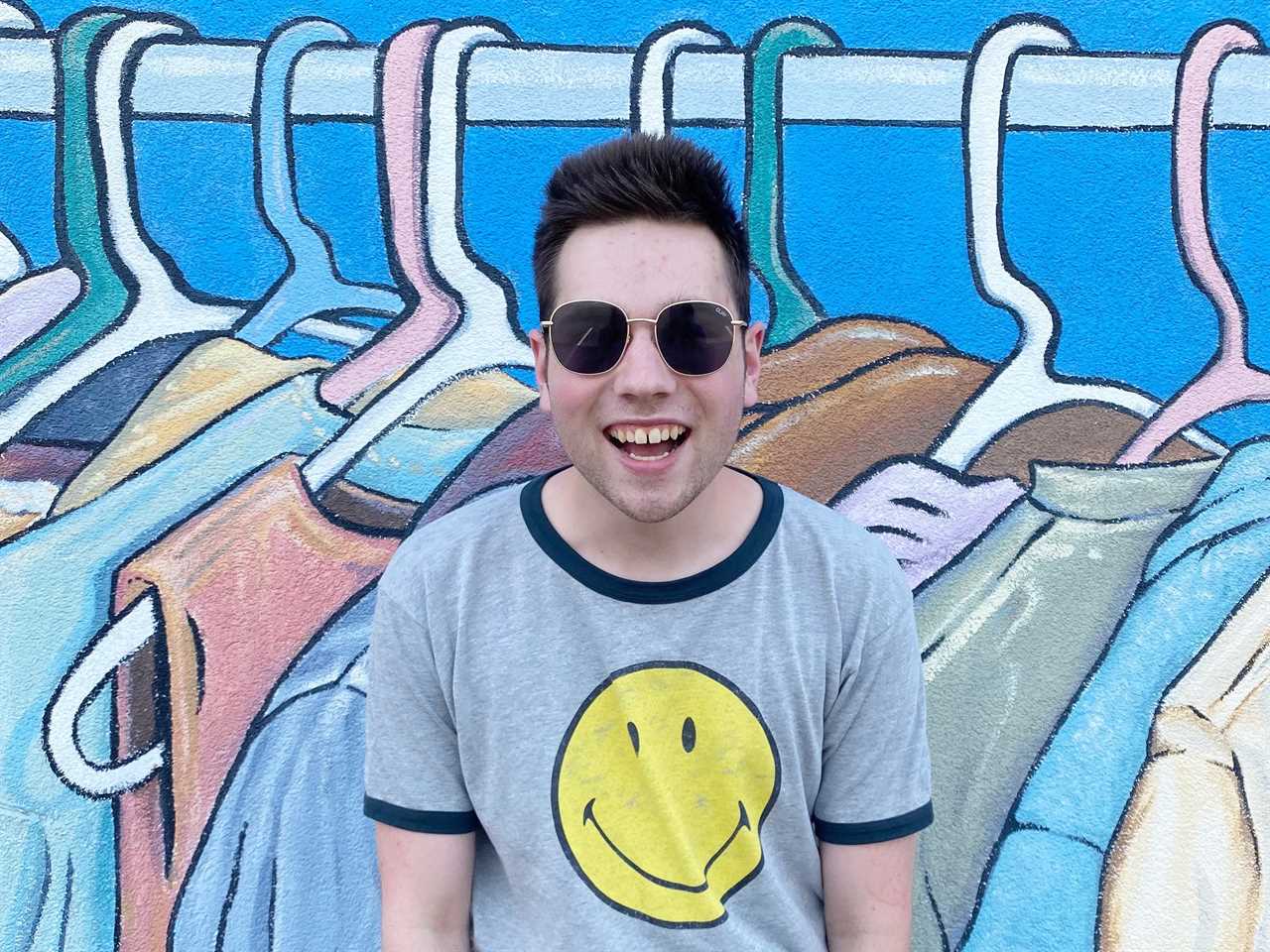 man smiles in front of a colorful mural of clothes hanging on a rack. he wears a smiley face shirt