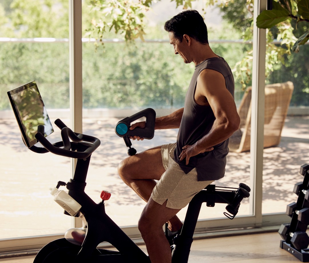 Caucasian man on Peloton stationary bike using massage gun on quad