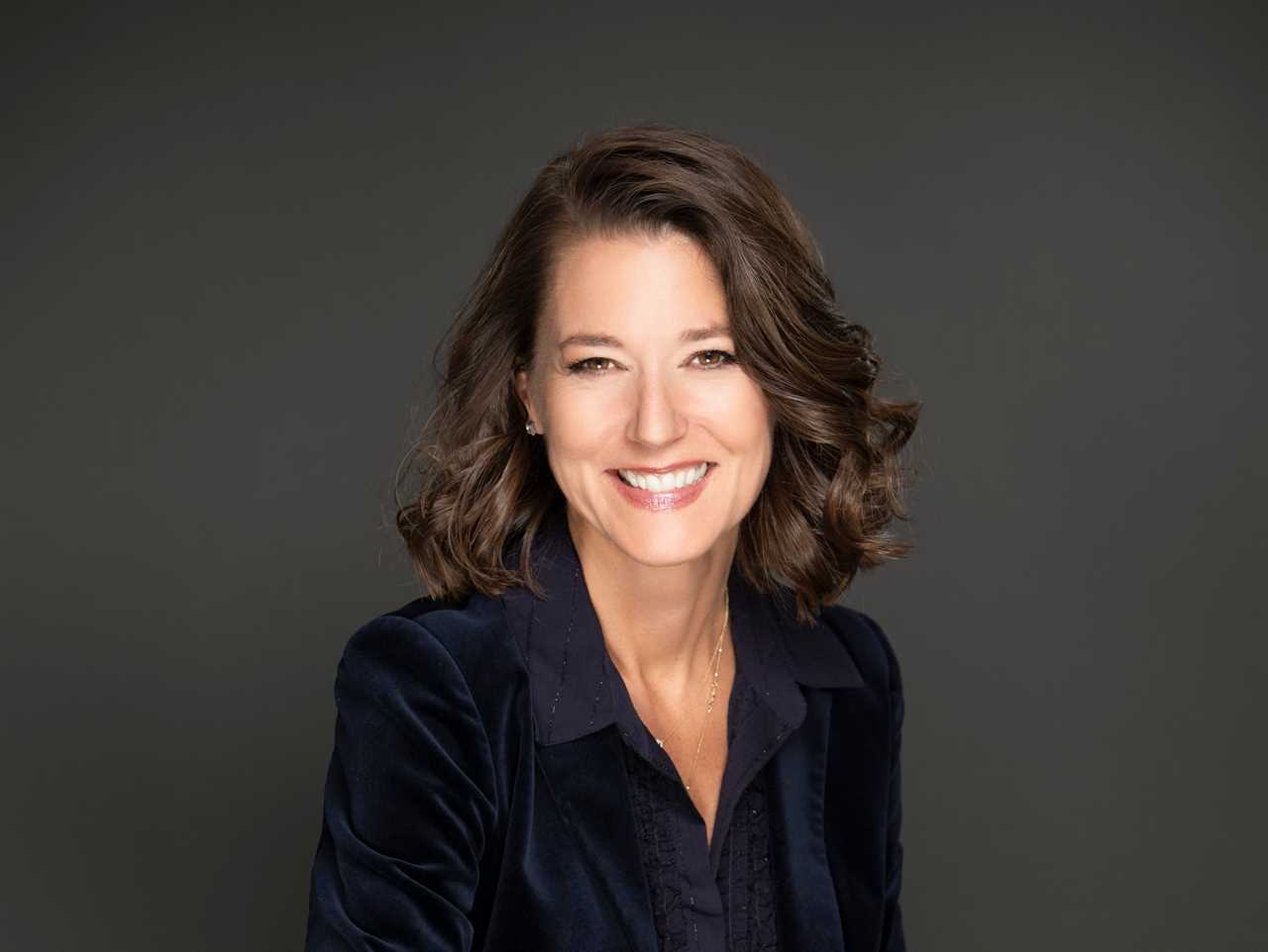 Mary Ellen Coe headshot, wearing black suit and sitting in a wooden chair