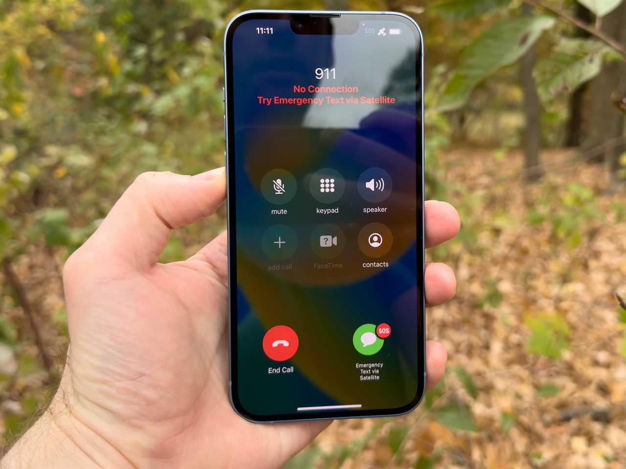 A hand holding an iPhone outside showing Apple Emergency SOS via Satellite.