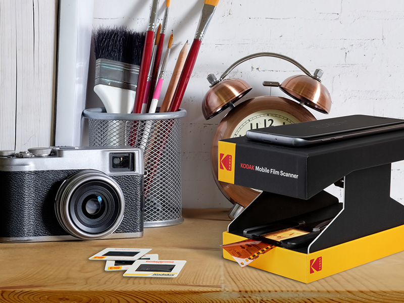 a Kodak Mobile Film Scanner sits on a desk with a camera, alarm clock and more