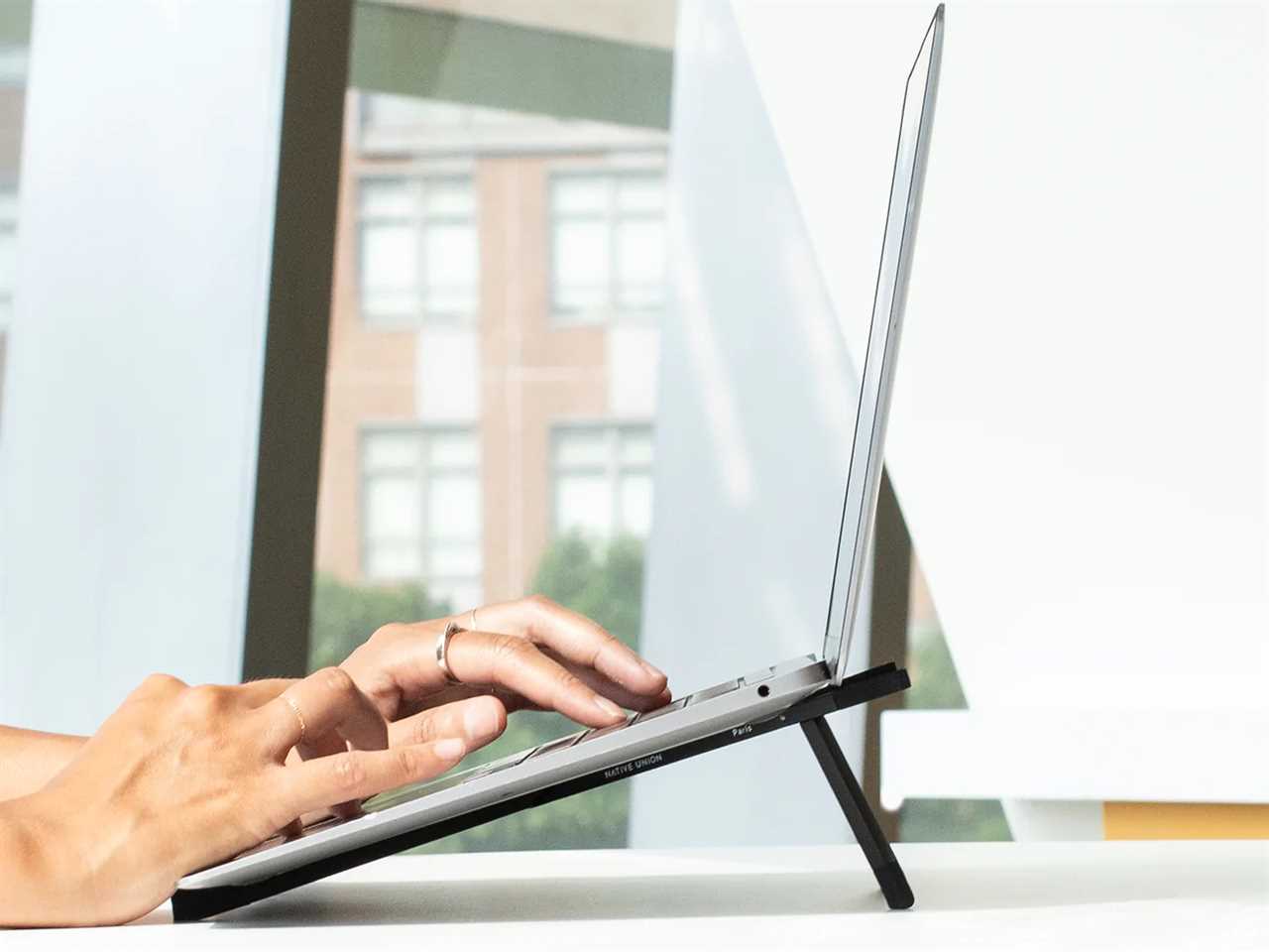 a person uses their laptop mounted on a Native Union Fold Laptop Stand with a window in the background as one of the best tech products under $50