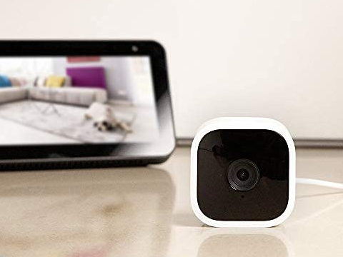 a Blink Mini Security Camera sits on a surface in front of a tablet as one of the best tech products under $50