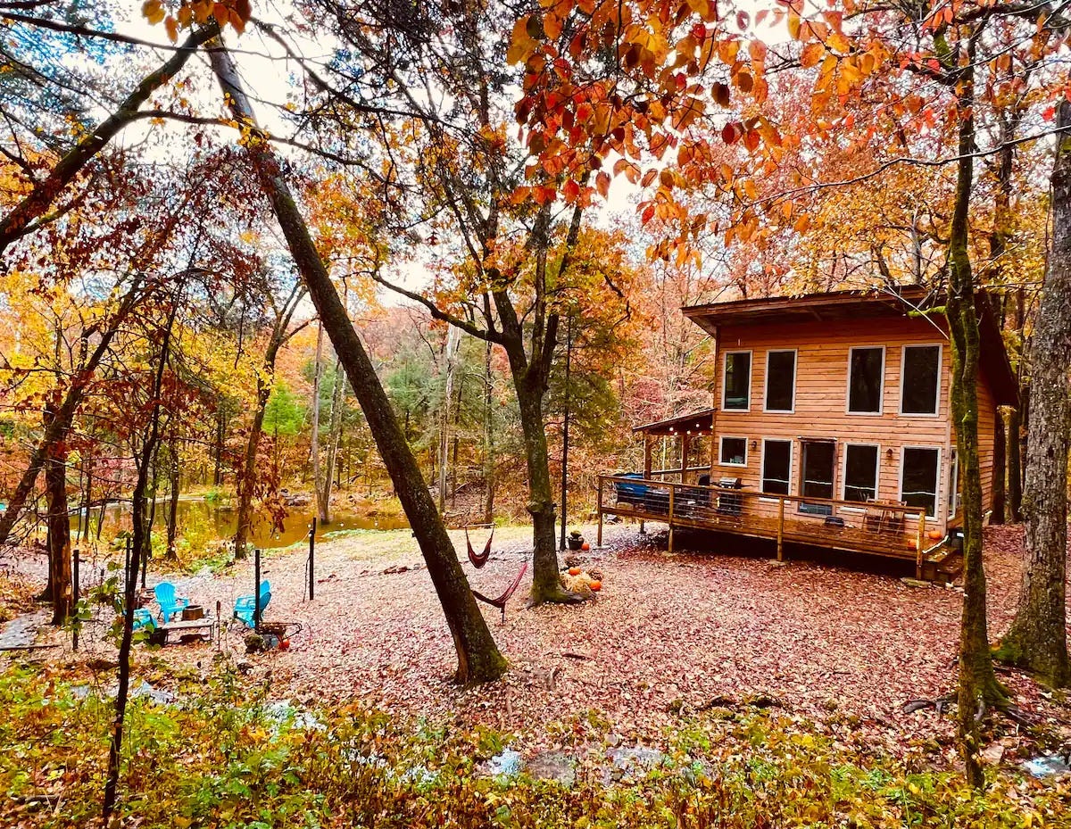 A wooden house surrounded by orange and red trees, piles of fallen leaves, and blue adirondack chairs around a campfire