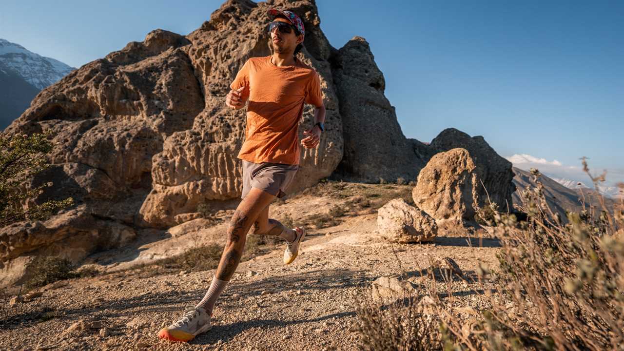Man in an orange shirt, cap, sunglasses, and hybrid running shoes running on a mountain trail with rocks in the background.