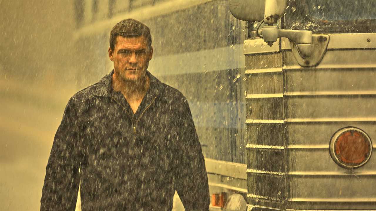 Actor Alan Ritchson walks outside through the rain beside a bus in the new Amazon series 