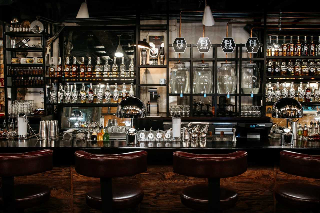 Interior of a darkly lit bar with lots of bottles of liquor, including aquavit, on the back wall with dark wooden bar stools in the foreground.