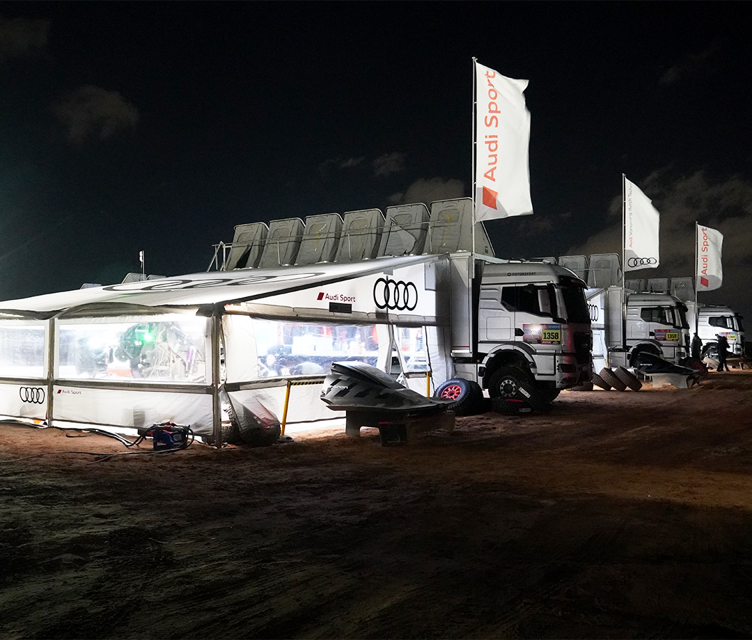 Dakar off-road big rigs lined up at night with lighted tents at night in the desert.