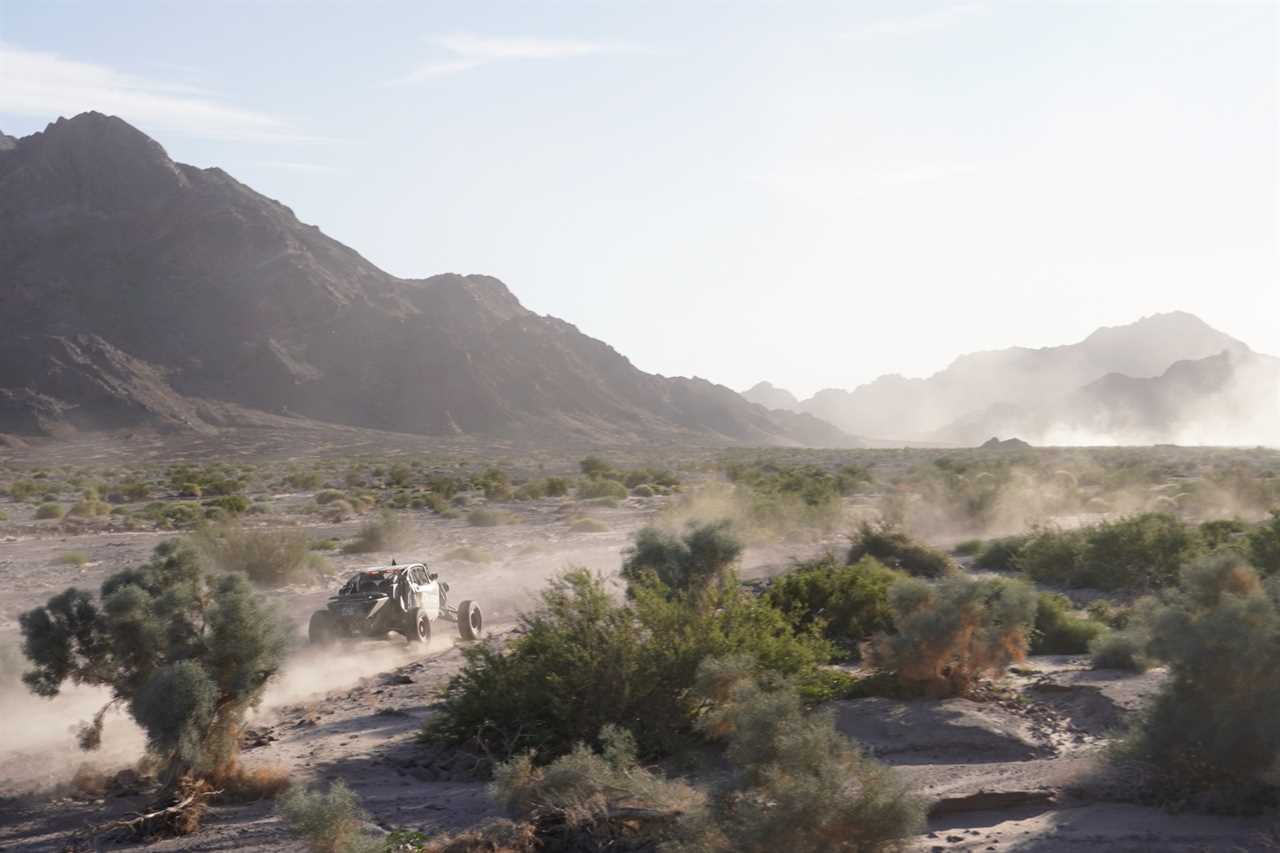 Dune buggy racing across a desert in the sunlight with mountains in the background.