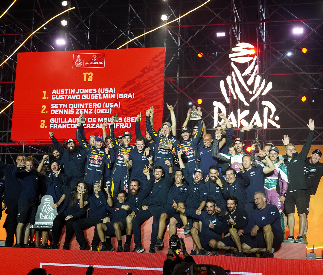 Group of Dakar Rally winners assembled in a group with the final leaderboard behind them.