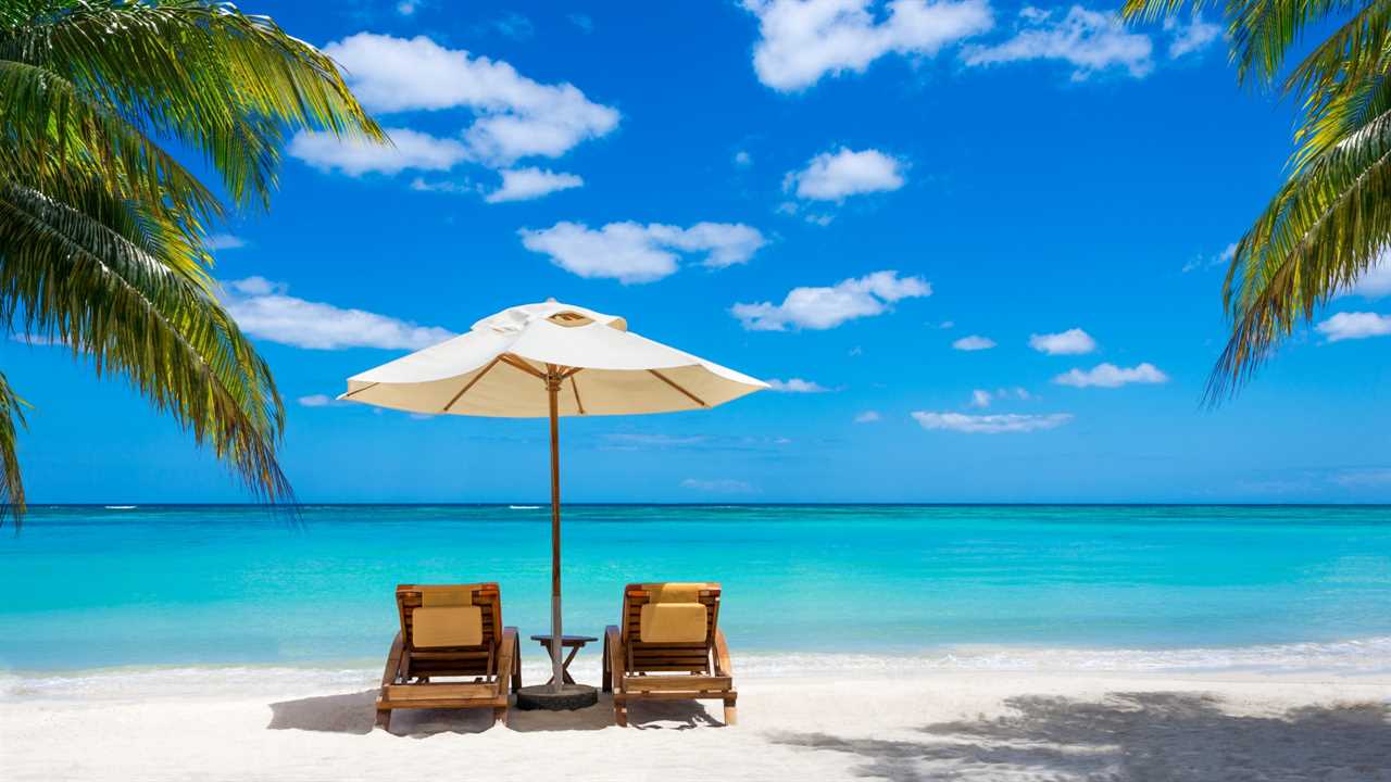 Two wooden deck chairs and a white umbrella on a tropical beach with the ocean beyond. winter getaway