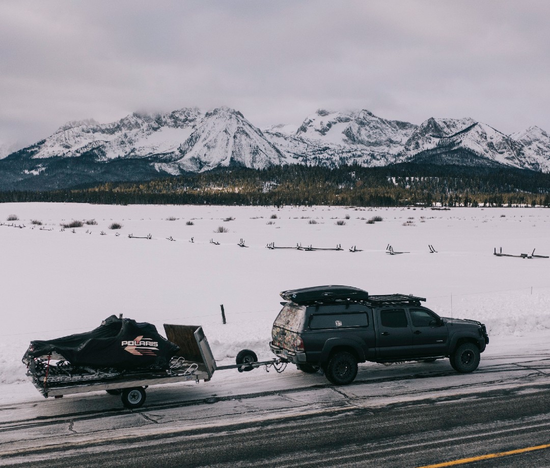 SUV pulling a trailer with a snowmobile on a highway in mountain country.