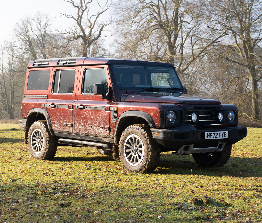 Muddy, boxy maroon 4x4 SUV parked in a field with trees in the background.