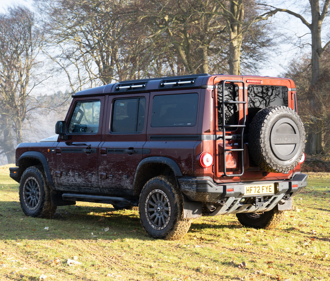 Rear shot of a boxy maroon 4x4 SUV that's muddy and parked in a field with trees in the background.