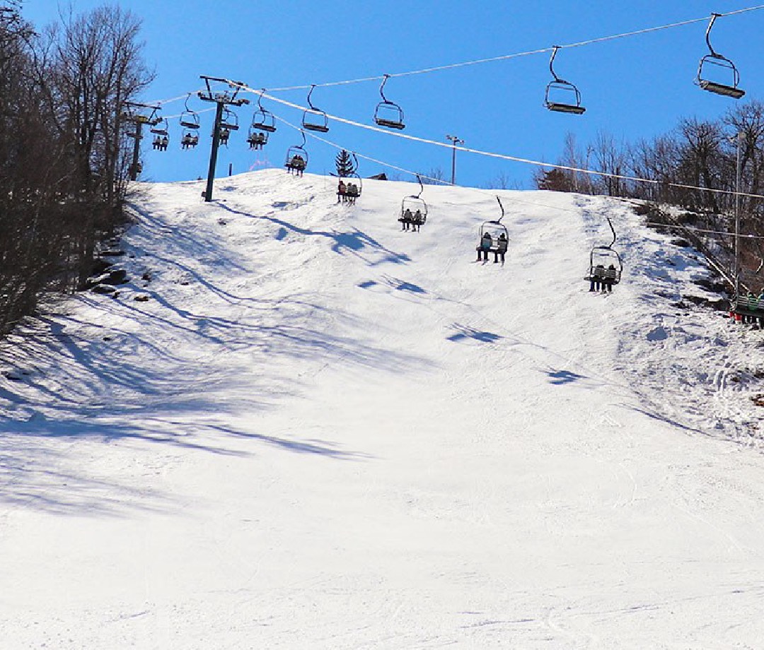 Ski slope with a chair lift with skiers in it going across with trees on either side.