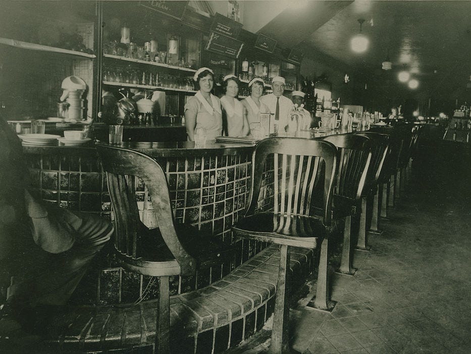 Black and white photograph of staff in uniform posed standing behind bar in 1924