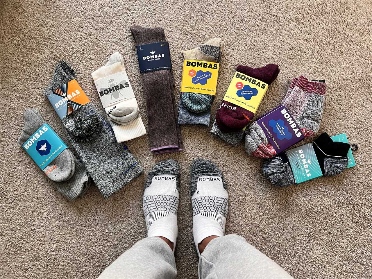 A person's sock-clad feet surrounded by a variety of Bombas socks on the ground.