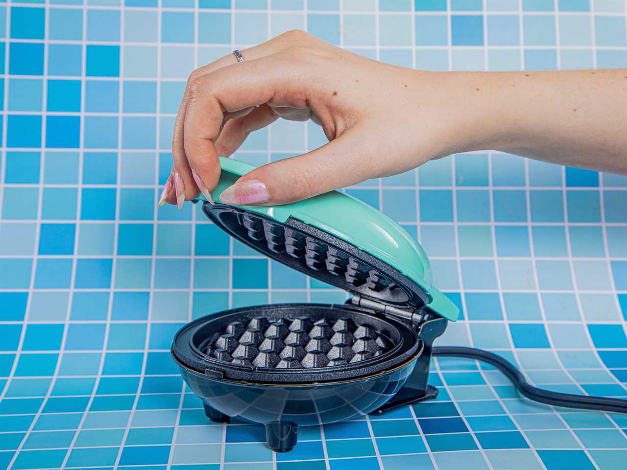 A teal Dash Waffle Maker being opened by a hand against a variety blue-toned tile background.