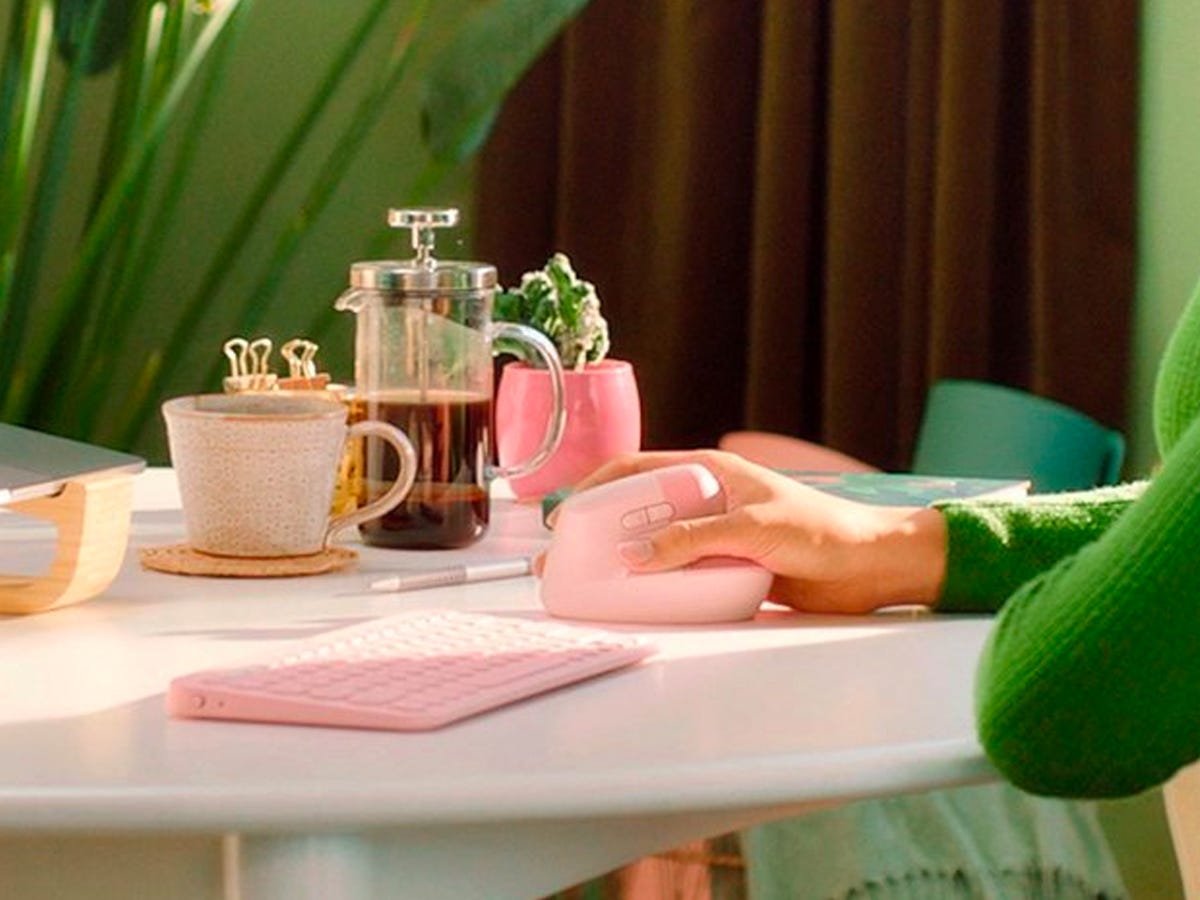 Person using a pink Logitech Lift Wireless Ergonomic Mouse at a desk with a matching keyboard, cup of coffee, and a small plant also on it.