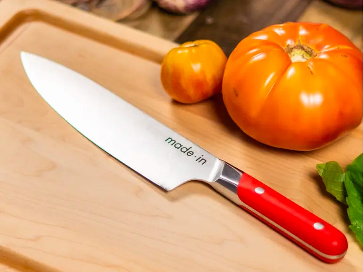 Made In 8 Inch Chef Knife with a red handle resting on a wooden cutting board next to two heirloom tomatoes.