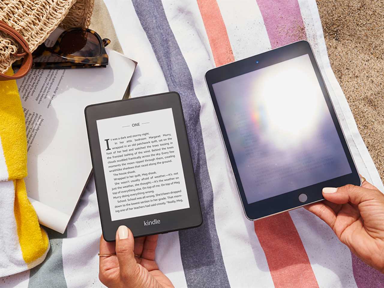Kindle Paperwhite displaying a book next to a tablet reflecting the sun at the beach.