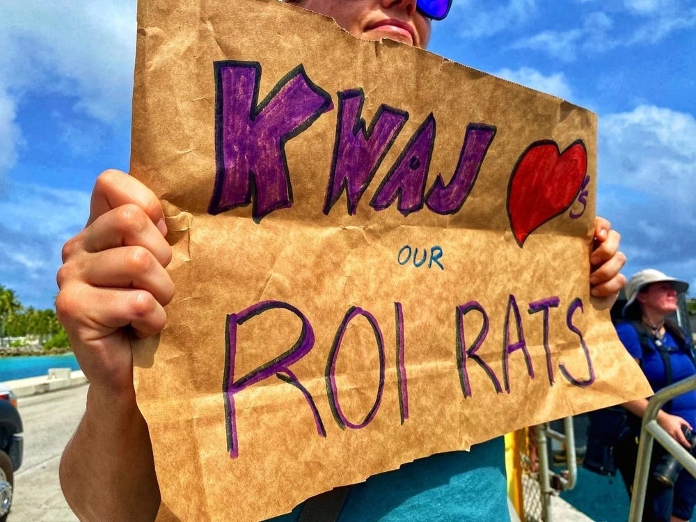 a woman holds a sign that says "Kwaj loves our Roi Rats," referring to residents of the Kwajalein Island and Roi-Namur Island, respectively