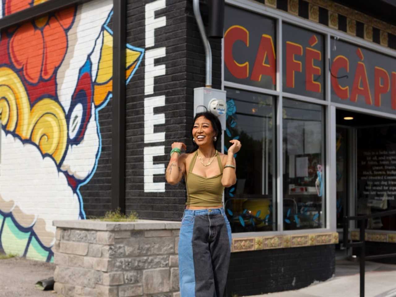 A woman wears an army green halter top and wide-leg jeans standing in front of a café that has colorful street art outside.