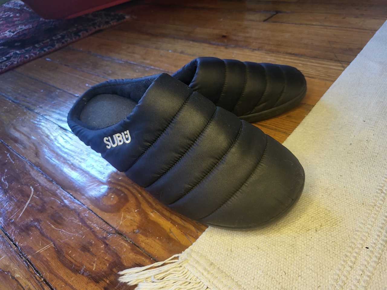 A pair of black Subu house slippers on a wooden floor with a rug.