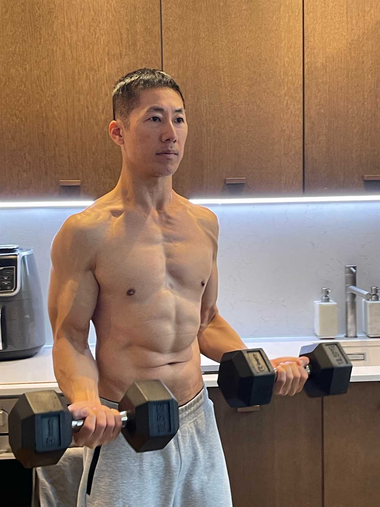 A shirtless, muscular man in sweatpants holding dumbbells.