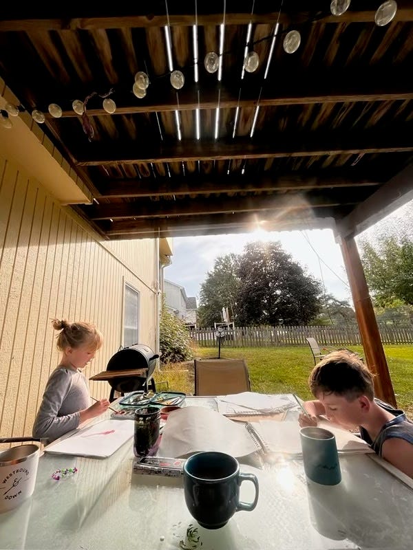 Two children draw on a patio table in a grassy backyard