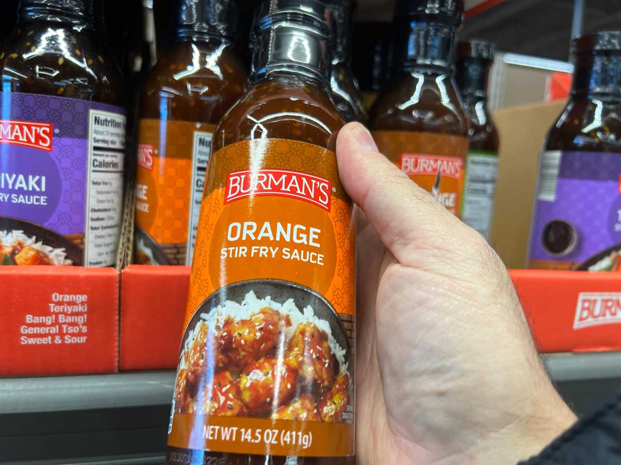 A hand holding a bottle of Burman's orange stir-fry sauce.