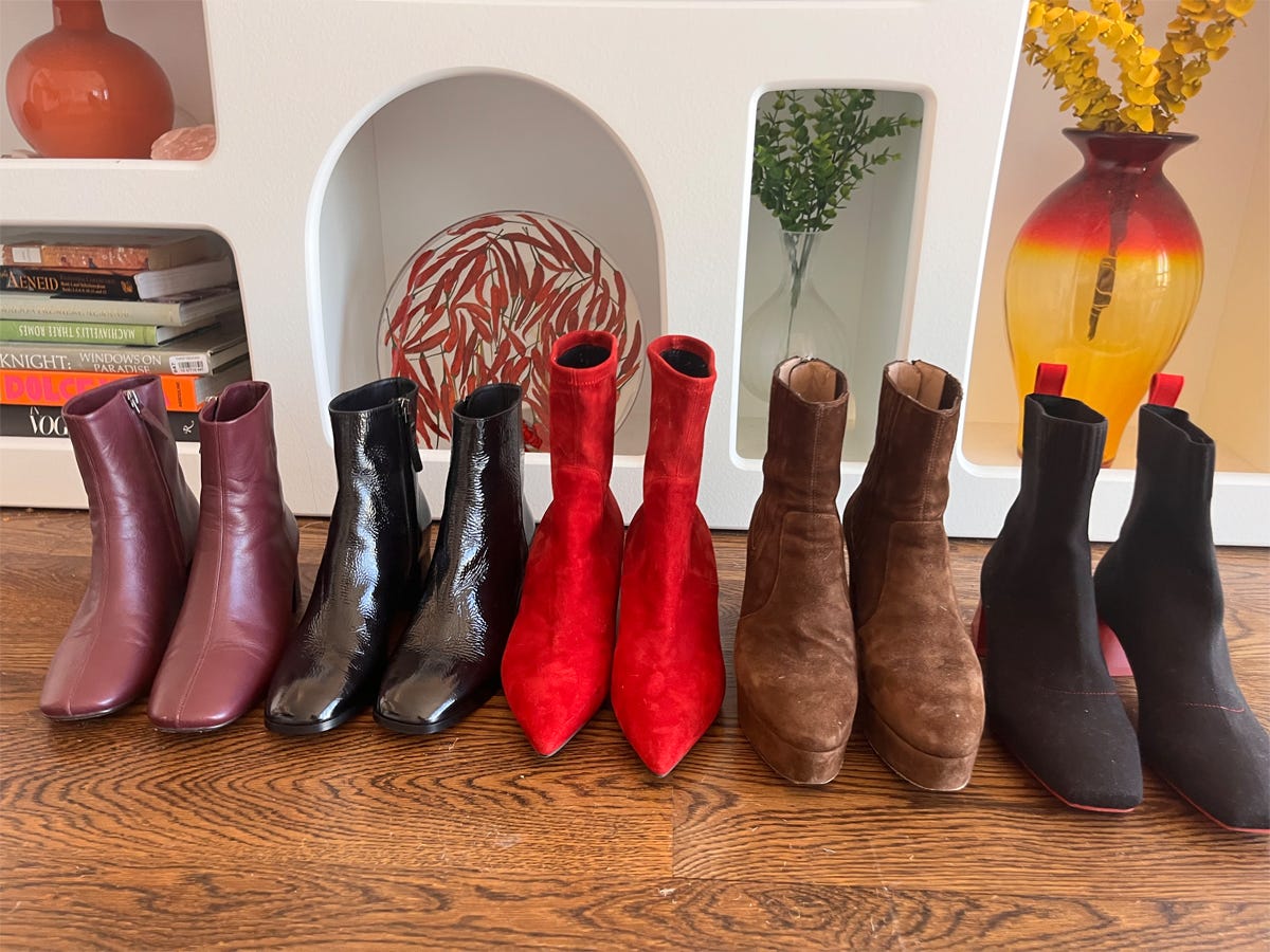 Assorted pairs of ankle boots in various colors set on a wooden floor in front of a bookshelf.