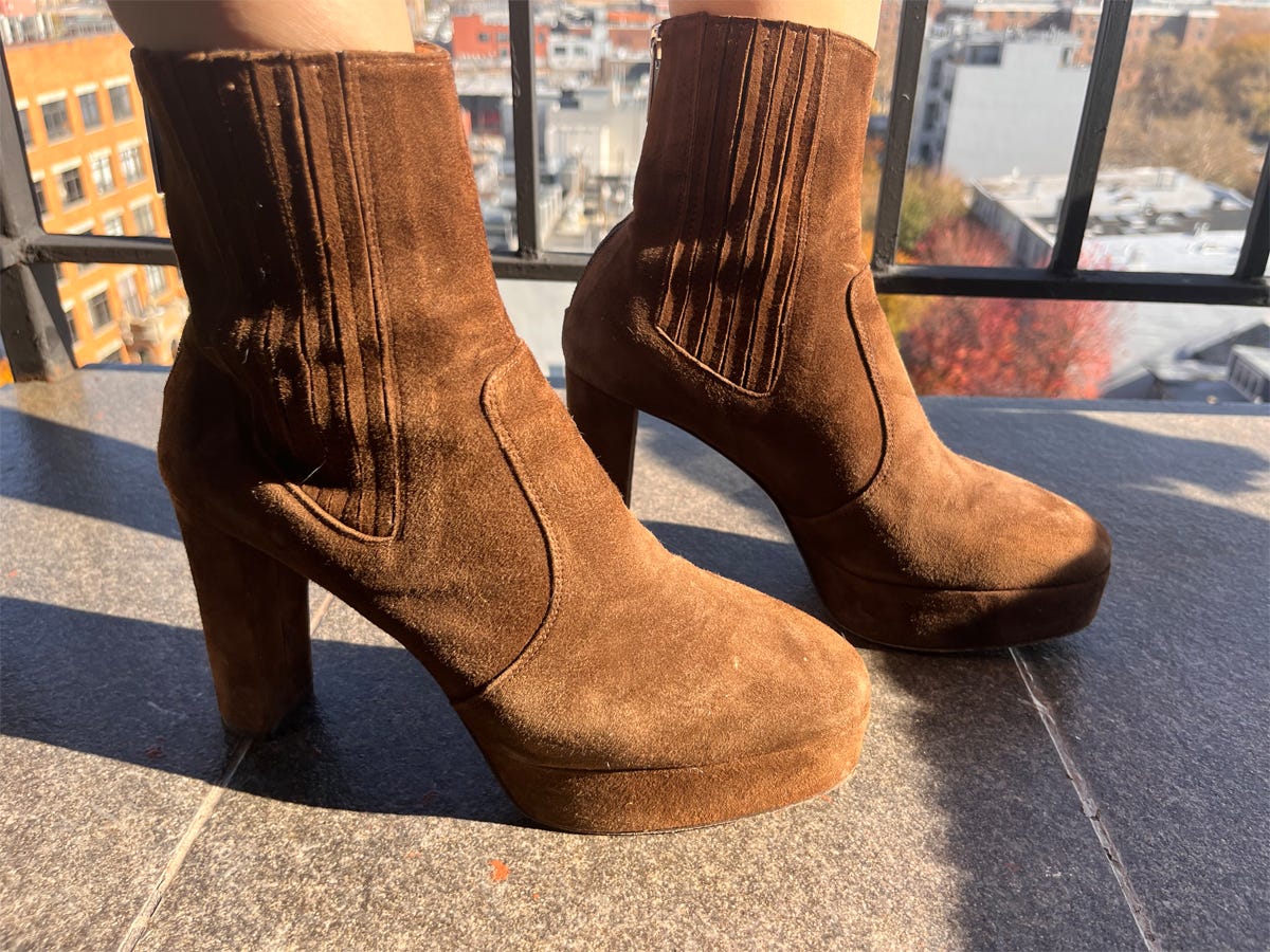Person's feet shown wearing INEZ Pilar platform ankle boots in brown suede while standing on a balcony.