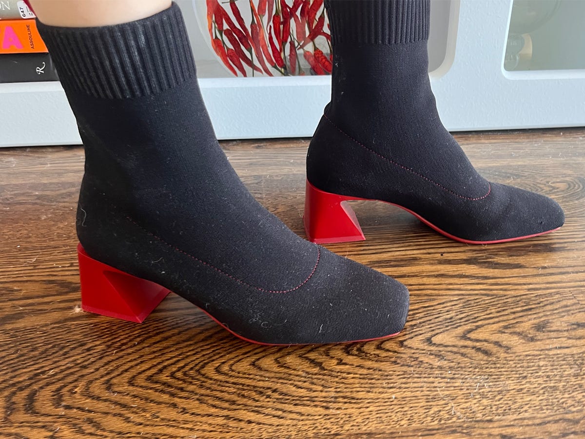 Person wearing square-toe black ankle boots with red heels while standing on a wooden floor.