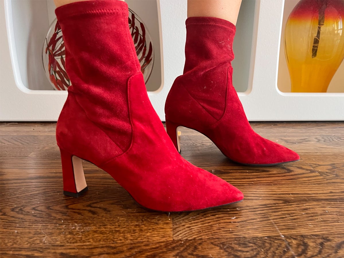 Person wearing a pair of red suede ankle boots while standing on a wooden floor in front of a bookshelf.