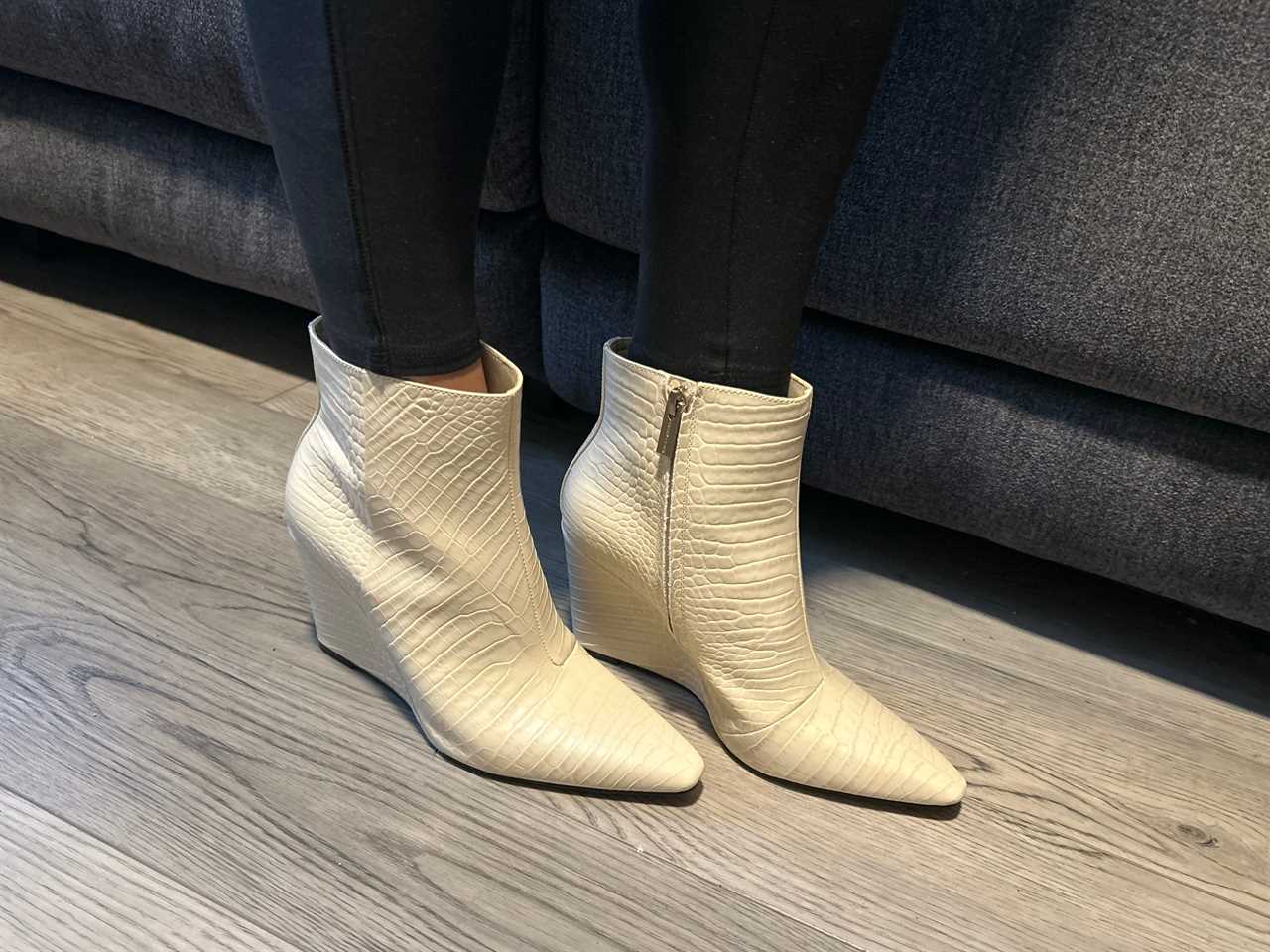 Person wearing a pair of white snakeskin wedge booties while standing in front of a couch.