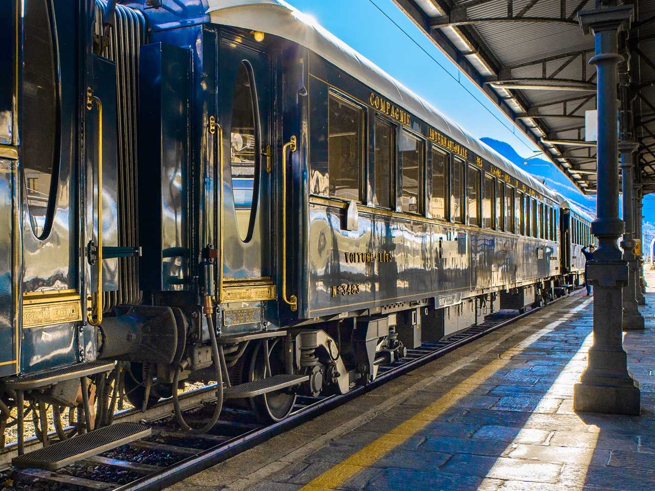 A navy blue train with gold trimmings stopped at a platform with mountains in the background.