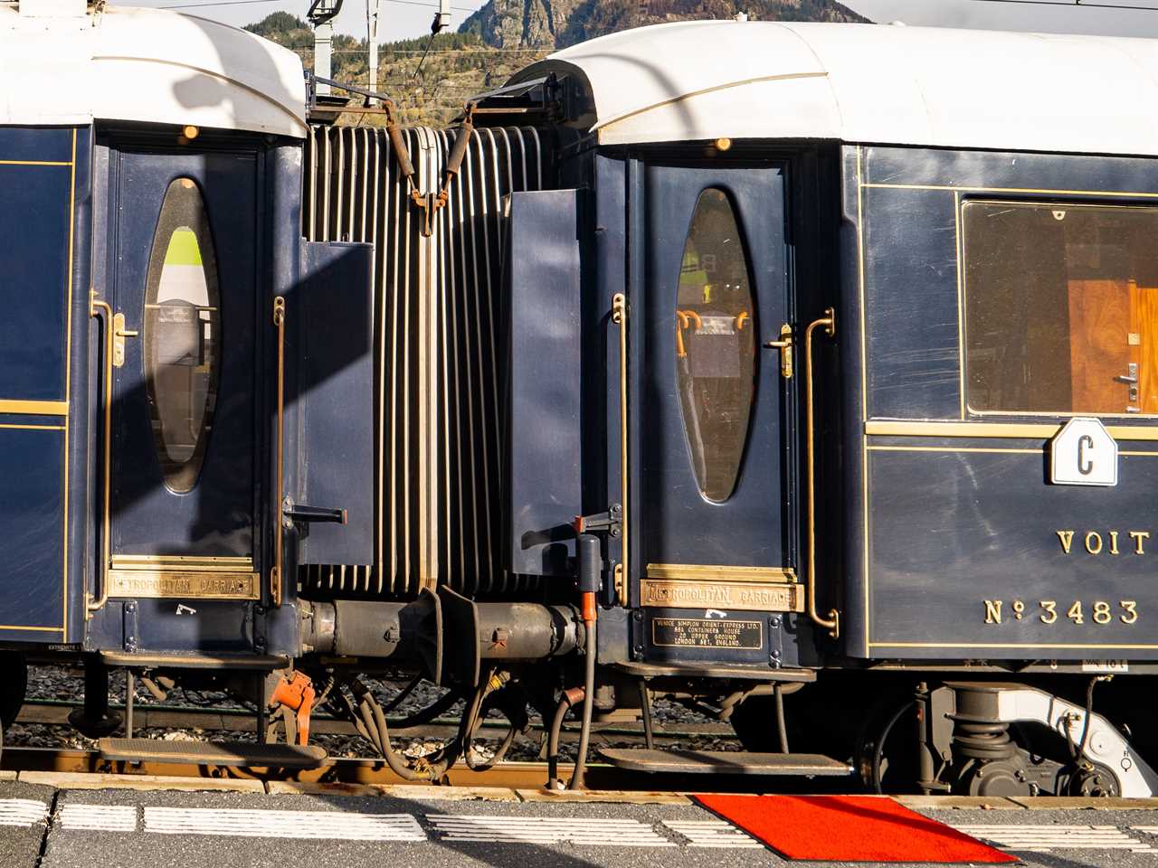 A navy blue train stopped at a platform with a red carpet in front of the door and mountains in the background